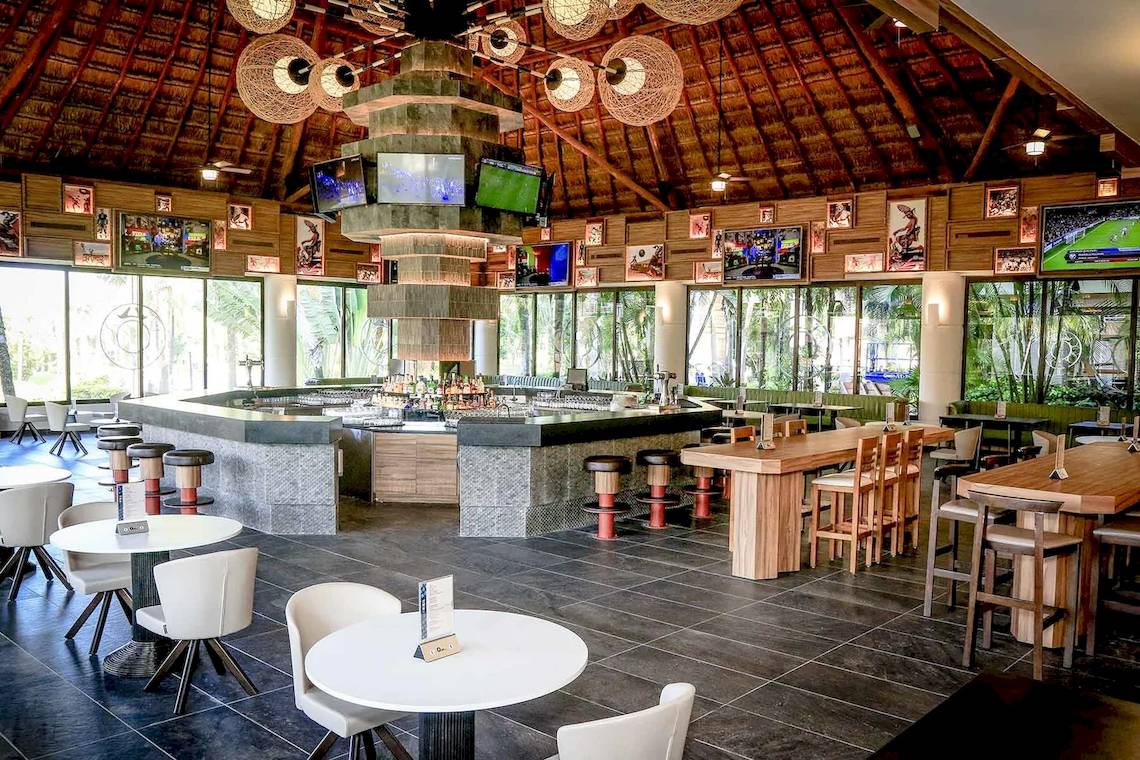 Innenansicht des Restaurants im Bahia Principe Luxury Akumal mit einer Bar, Tischen, Stühlen und Fernsehern an der Wand.