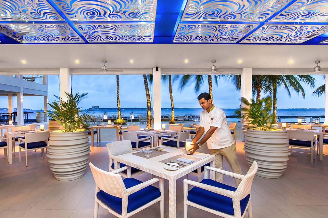 Innenansicht des Kurumba Maldives Restaurants mit Blick auf den Ozean und einem Kellner, der einen Tisch bedient.