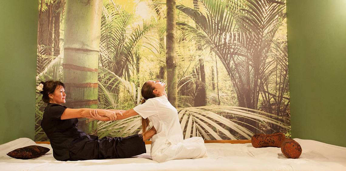 Zwei Frauen praktizieren Yoga in einem Hotel-Spa-Raum mit grüner Wand und tropischem Fototapete.