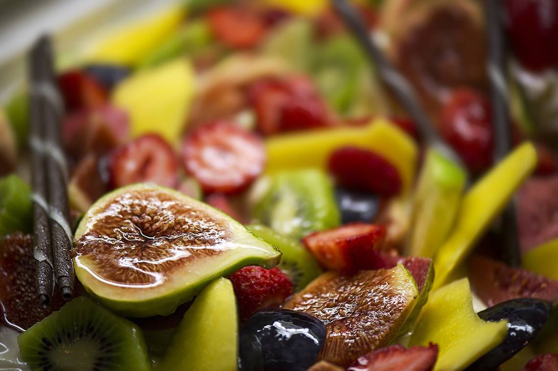 Nahaufnahme eines Obstsalats mit Feigen, Erdbeeren, Kiwis, Bananen und Trauben, mit einigen dunklen Schokostaebchen.