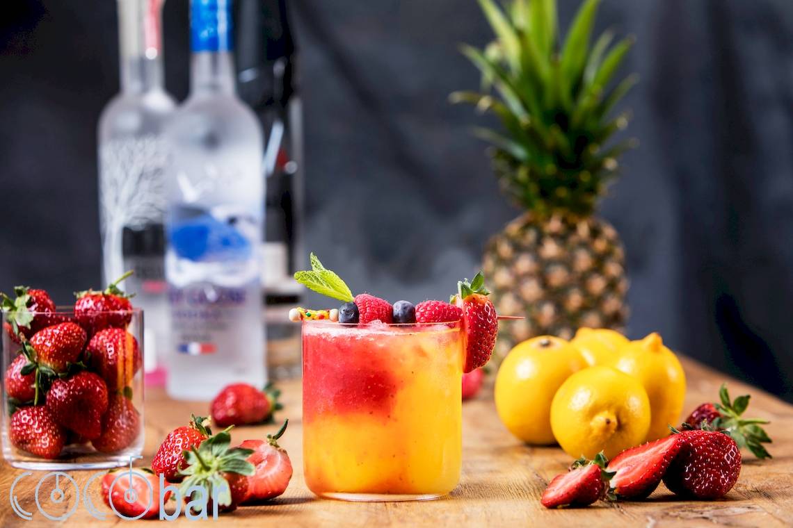 Ein Glas mit fruchtigem Cocktail, dekoriert mit Beeren und Zitrone, vor einem Hintergrund von weiteren Früchten und Vodka-Flaschen.