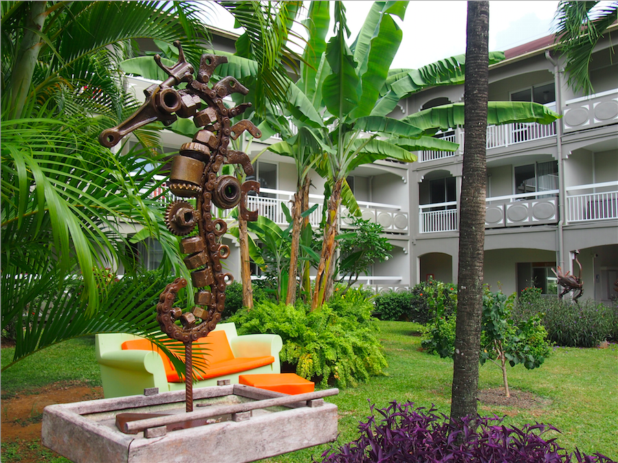 Tropischer Garten vor dem La Pagerie Hotel mit einer Skulptur, einem grünen Sofa und Palmen.