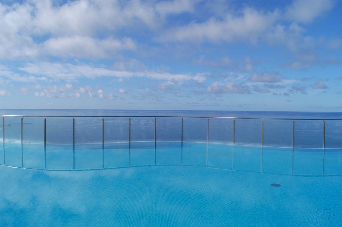 Ein blauer Pool mit Glasbarrieren bietet Aussicht auf das Meer und den Himmel mit Wolken.