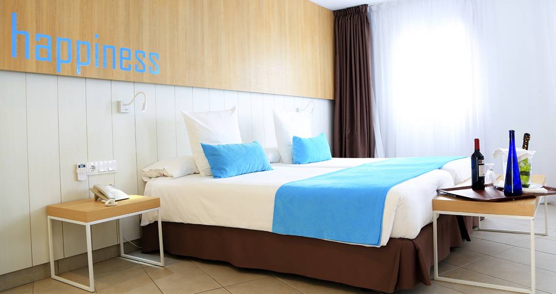 Zimmer im Labranda Playa Bonita Hotel mit zwei Betten, blauen Kissen und einer Tafel mit Weinflaschen.