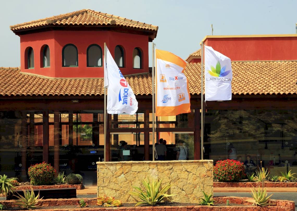 Eingang des Bluesea Aloe Corralejo Hotels mit bunten Flaggen und Gebäuden im Hintergrund.