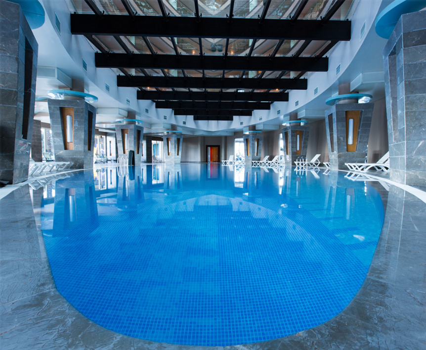 Innenpool des Long Beach Alanya Hotels mit weißen Liegestühlen und einer Glasdecke.