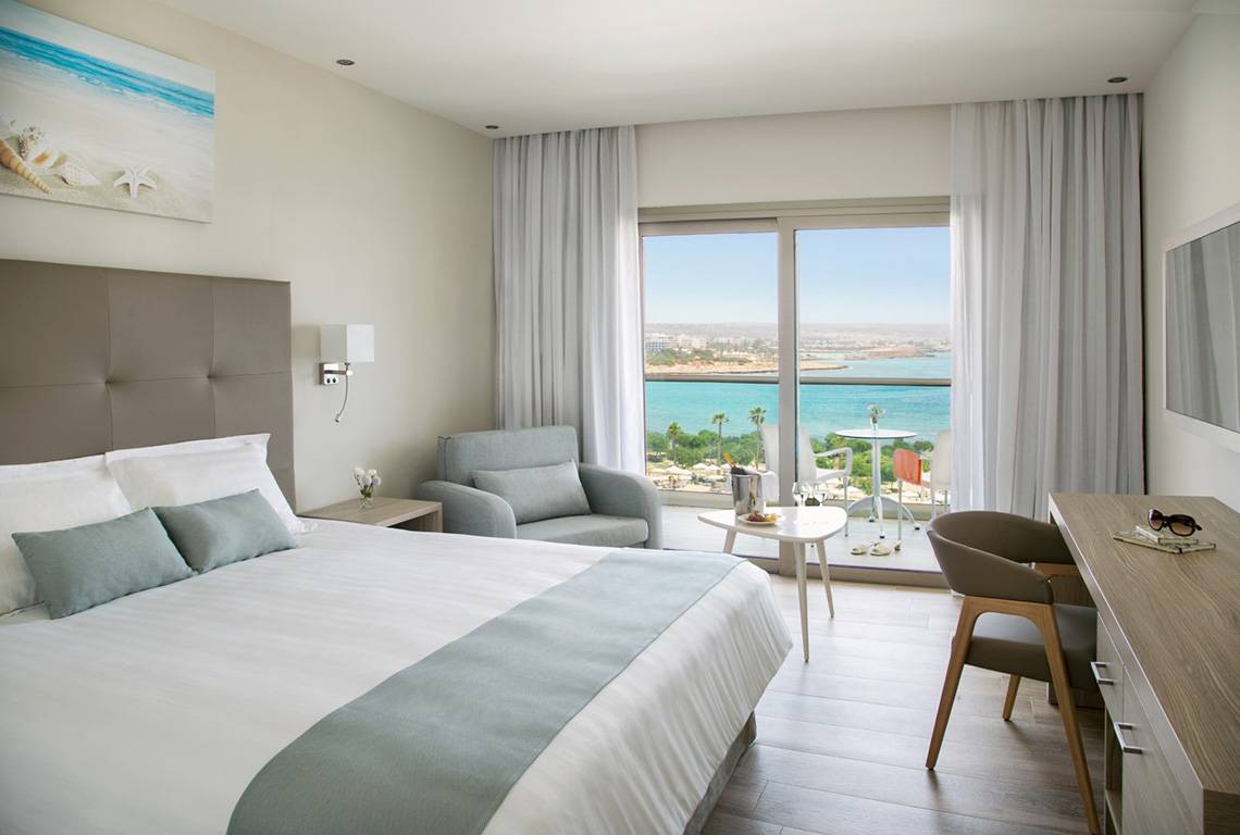 Interieur eines Hotelzimmers im Asterias Beach Hotel mit Blick auf das Meer und eine Terrasse.