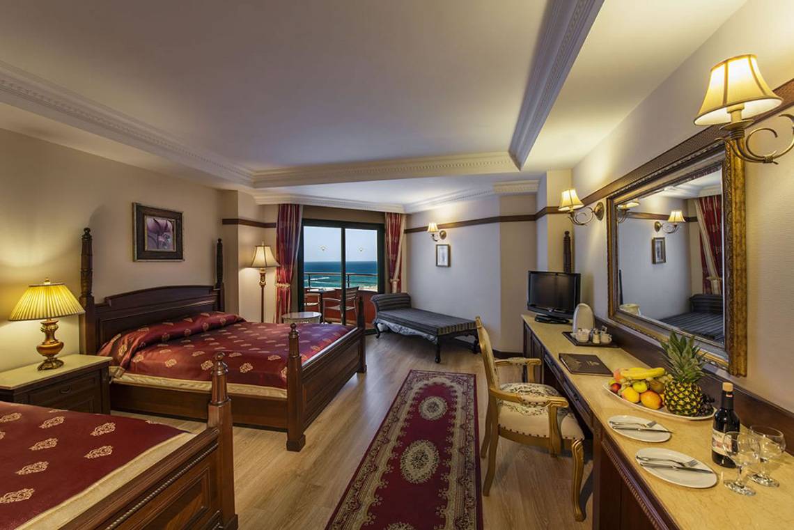 Innenansicht eines Hotelzimmers im Delphin Palace Hotel mit zwei Betten, einem Schreibtisch, einem großen Spiegel und einem Fenster mit Meerblick.