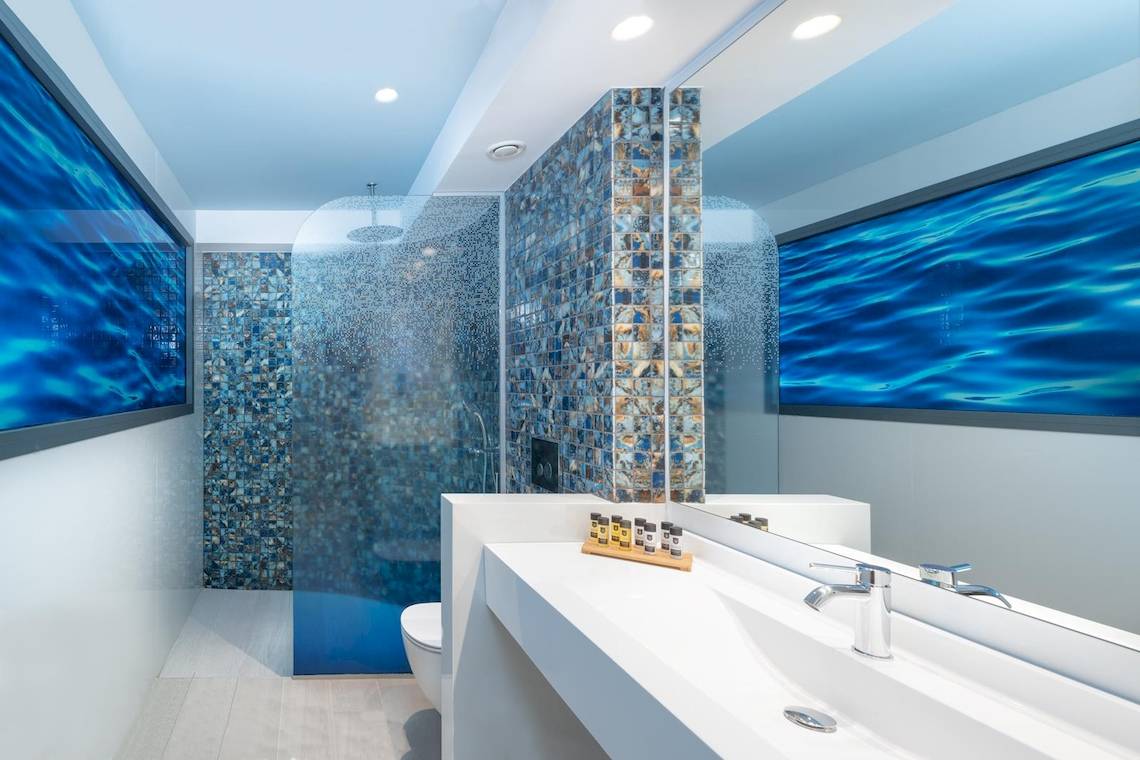 Das Badezimmer des Lyttos Mare Hotels mit blauen Fliesen, einer Regendusche und einem großen Spiegel.