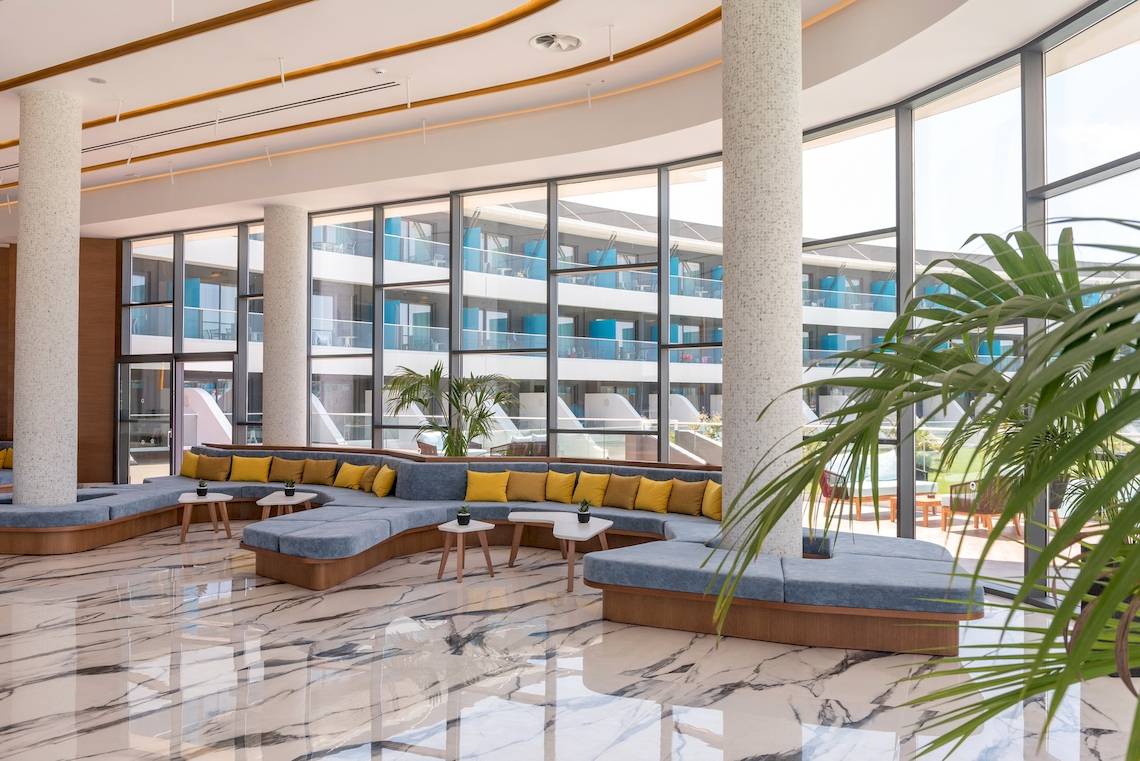 Lobbybereich des Lyttos Mare Hotels mit blauen Sofas, gelben Kissen und großen Fenstern.