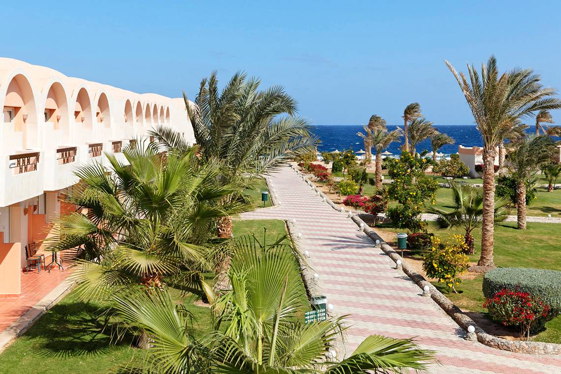 Außenbereich des Three Corners Sea Beach Resorts mit Gehweg, Palmen und Meer im Hintergrund.