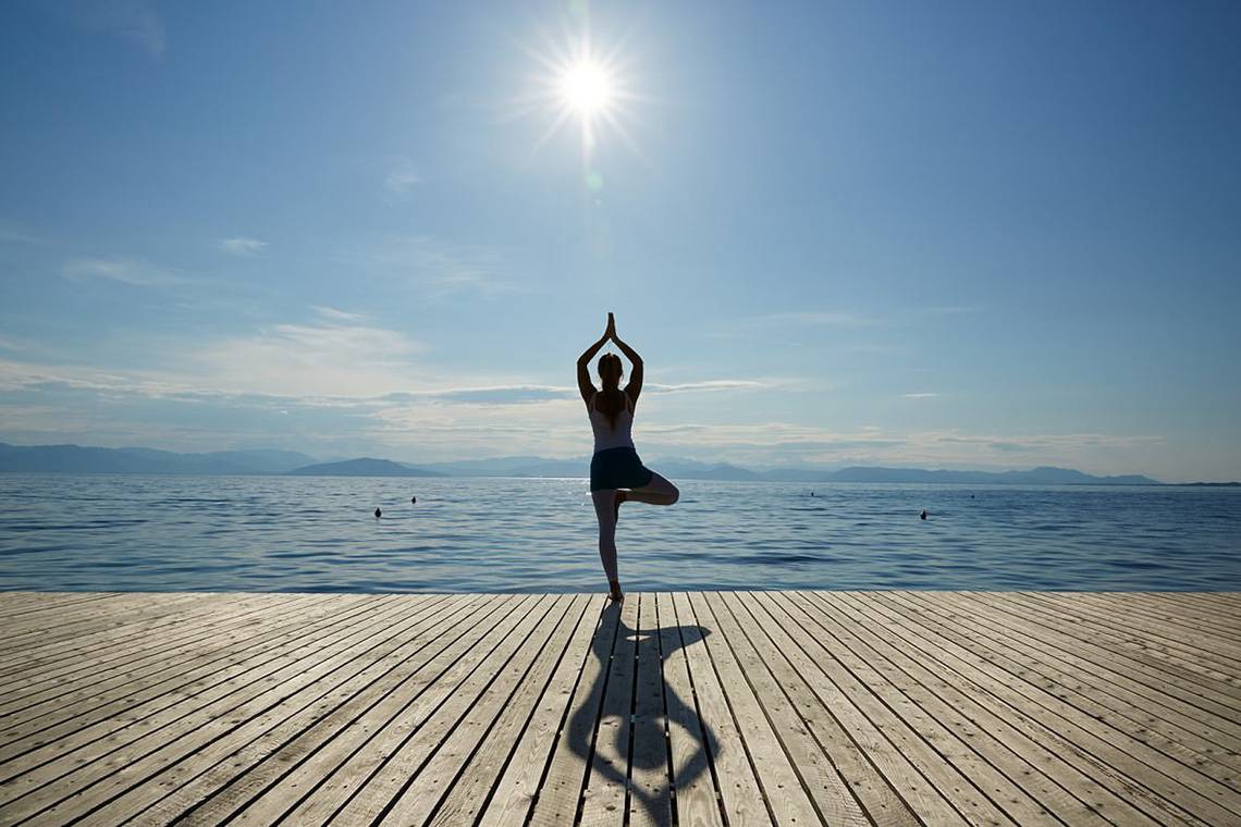 Eine Person praktiziert Yoga in einer Balance-Pose auf einer Holzplattform am Meer.