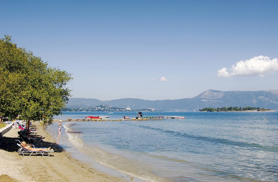 Strandszene am Louis Kerkyra Blue Hotel mit Liegestühlen, Bäumen und Bergen im Hintergrund.