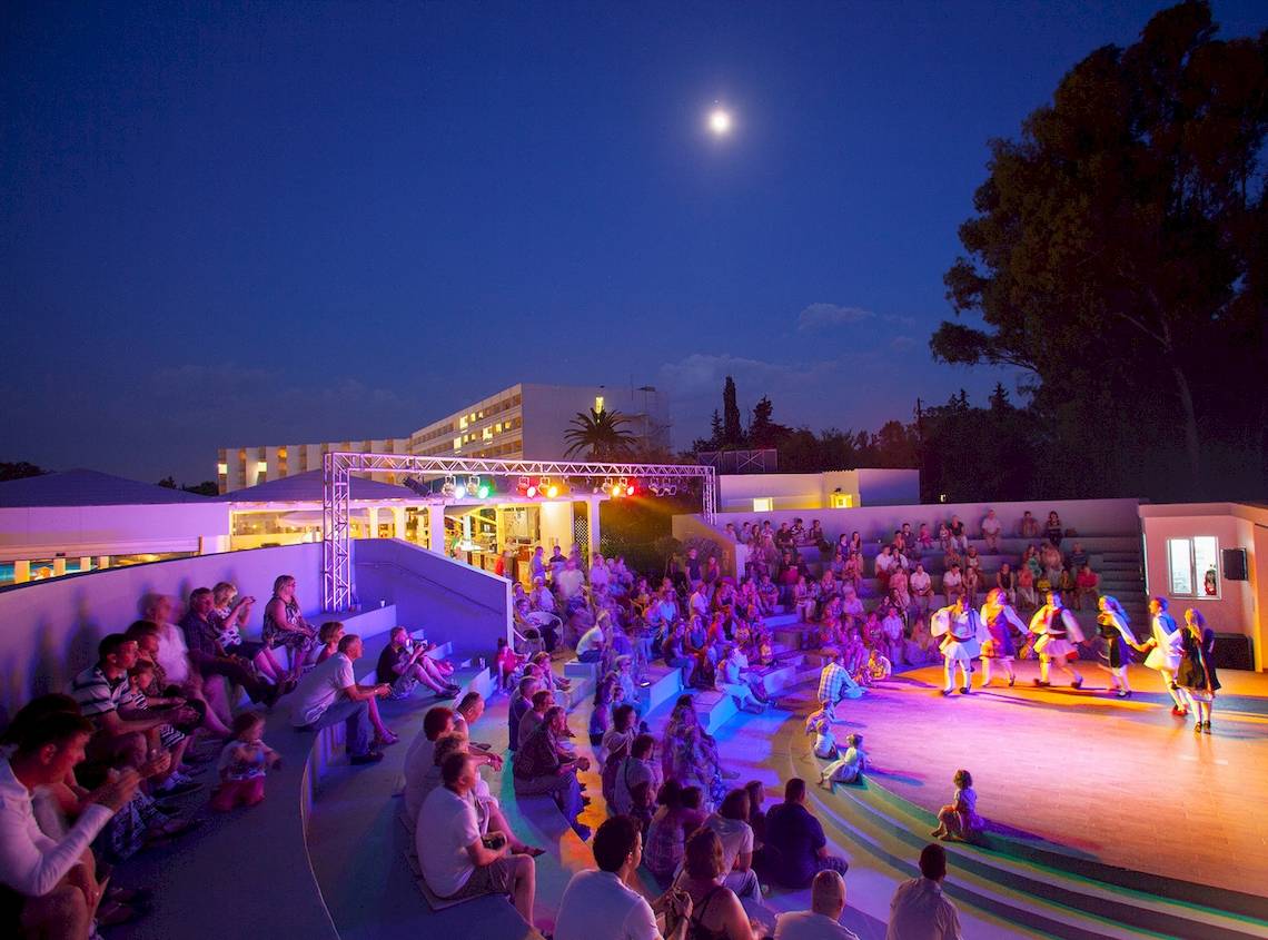 Menschen sitzen auf Stufen und beobachten eine Aufführung auf einer Bühne, Louis Kerkyra Blue Hotel bei Nacht.