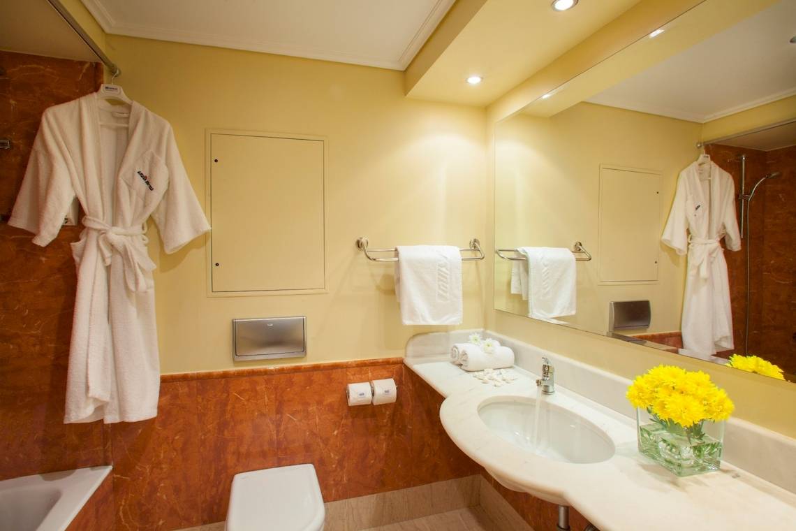 Badezimmer im Louis Kerkyra Blue Hotel mit Badewanne, Toilette, Waschbecken und gelben Blumen.