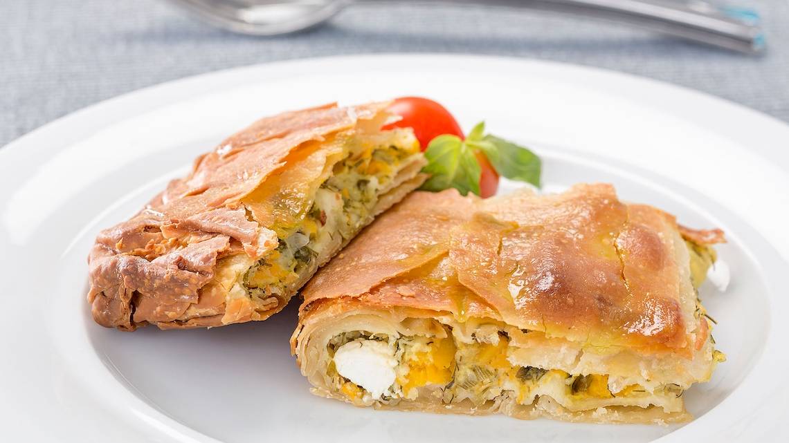Zwei Stücke Spinat und Feta in Filoteig auf einem weißen Teller mit einer Tomate und Basilikum.
