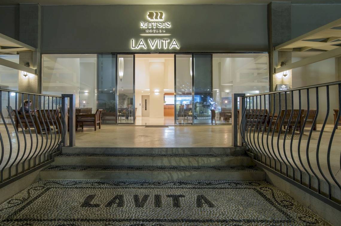 Außenansicht des Mitsis La Vita Beach Hotels mit beleuchtetem Logo und Eingangsbereich.