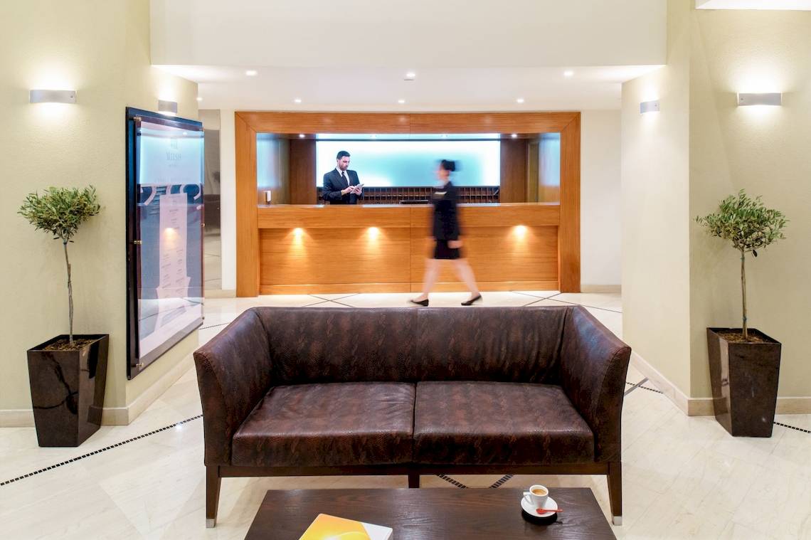 Hotelrezeption mit Sofa und Pflanzen, Mann am Empfang und Frau in Bewegung.