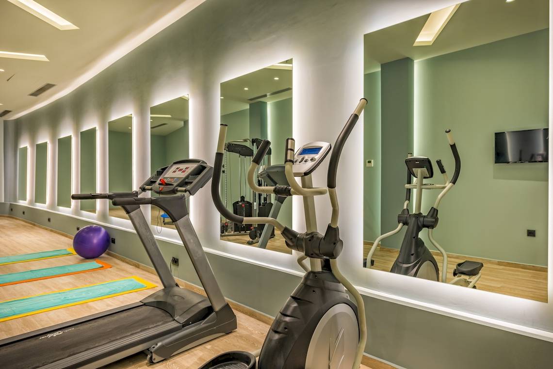 Fitnessstudio mit Laufband und elliptischer Maschine, Spiegelwänden und einer Yogamatte.