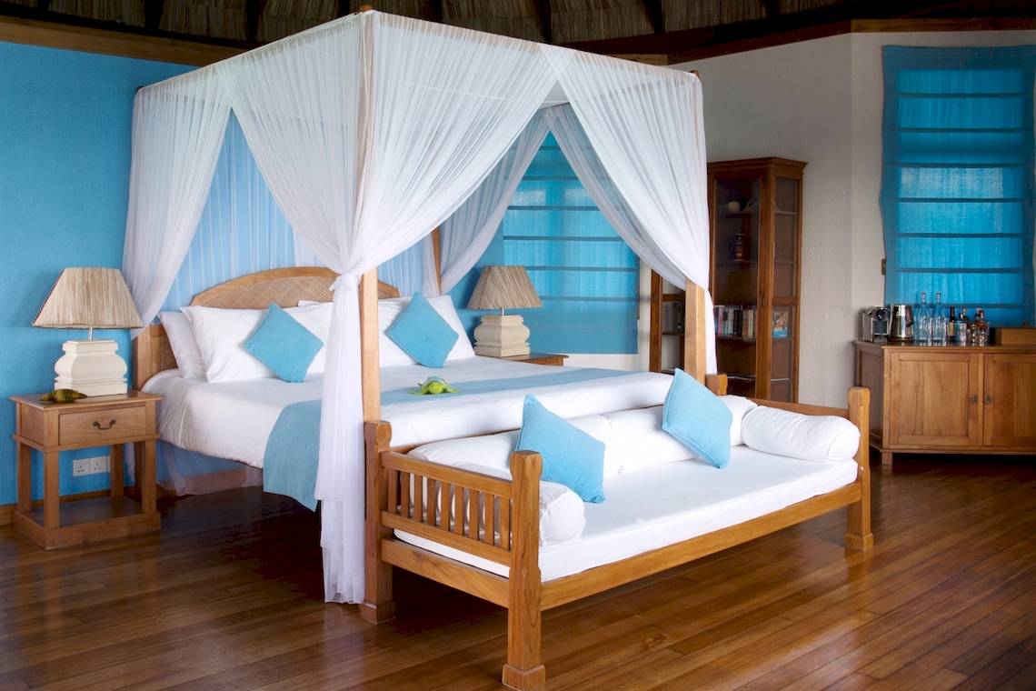 Coco Palm Dhuni Kolhu Schlafzimmer mit Himmelbett, blauen Kissen und Holzmöbeln