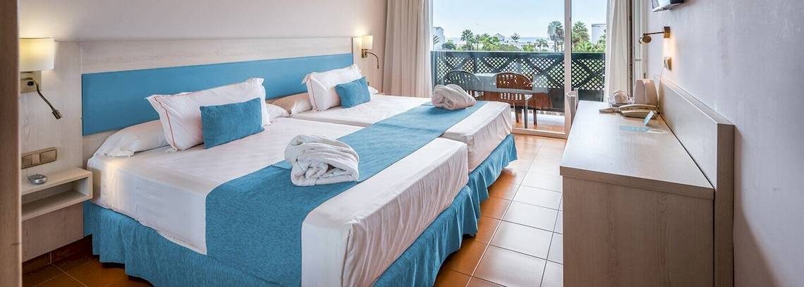 Doppelzimmer im Bluesea Costa Bastian Hotel mit zwei Betten, blauen Kissen, einer Nachttischlampe und einem Balkon.