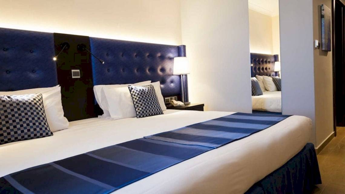 Ein geräumiges Hotelzimmer mit blauen Kopfteilen und weißen Kissen, AX The Victoria Hotel.