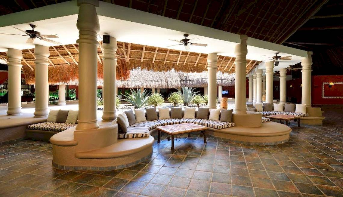 Innenbereich des Hotels Catalonia Playa Maroma mit Sitzbereichen, Tischen und Deckenventilatoren.