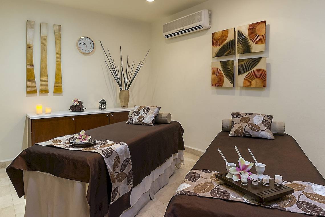 Massage-Behandlungsraum im Ocean Maya Royale mit zwei Massageliegen, dekorativen Elementen und einer Uhr an der Wand.