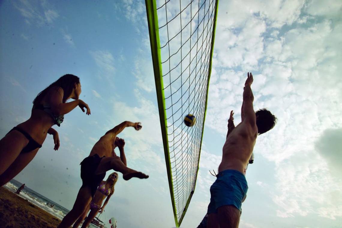 Mehrere Personen spielen Volleyball auf einem Strand, einer Spieler springt um das Netz.