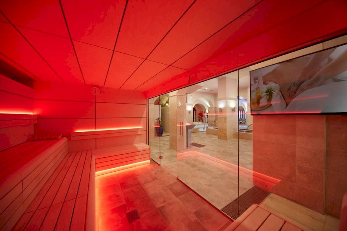 Ein rot beleuchteter Saunabereich im Secrets Lanzarote Resort & Spa mit Holzbänken und einer Glaswand.