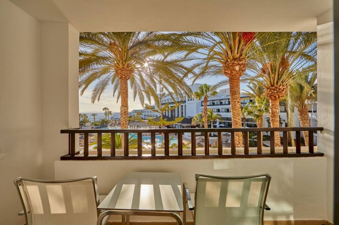 Balkon mit Tisch und Stühlen, Ausblick auf Palmen und ein Hotelgebäude.