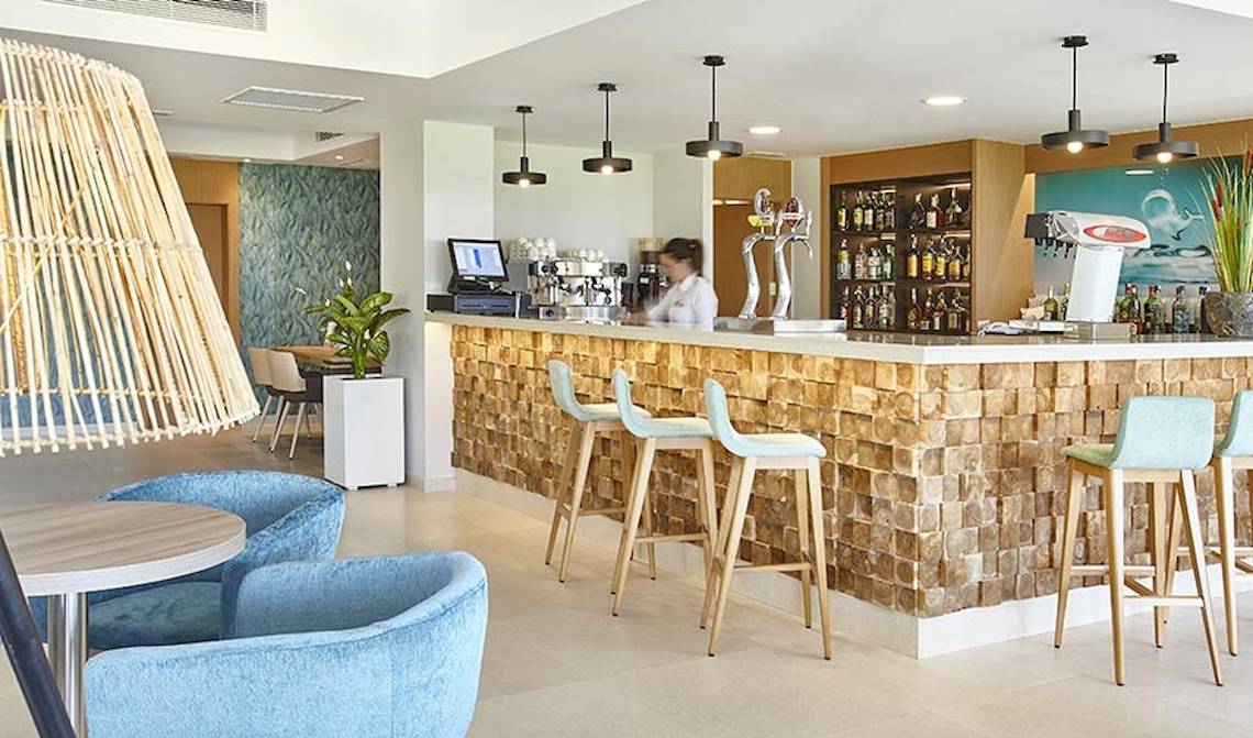 Innenbereich der Bar im Castell Royal Hotel mit Holztheke, blauen Stühlen und einer Angestellten.