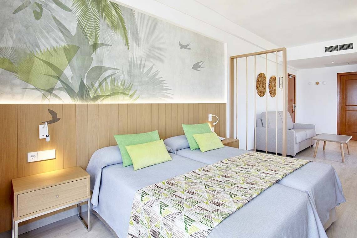 Schlafzimmer im Castell Royal Hotel mit zwei Betten, grünen Kissen und tropischem Wandbild.