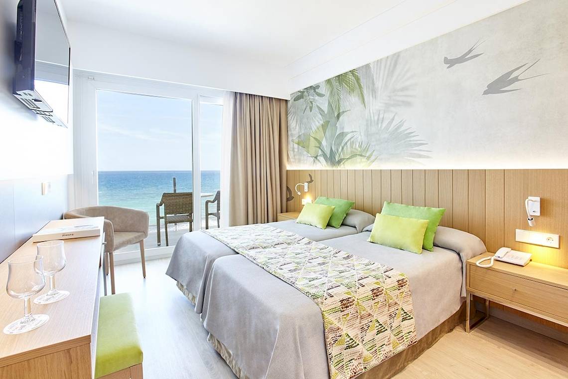 Zimmer im Castell Royal Hotel mit Blick auf das Meer, zwei Betten, grünen Kissen und einem Fernseher an der Wand.