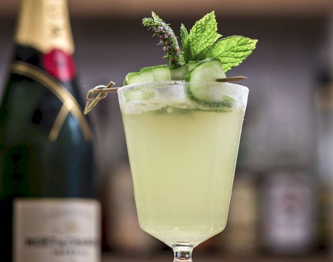 Ein Glas mit einem Cocktail, garniert mit Gurkenscheiben und Minze, vor einer Flasche Champagner.
