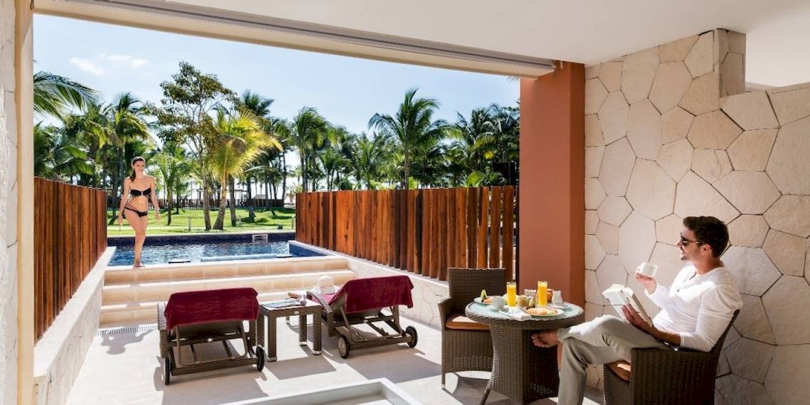 Ein Mann liest auf der Terrasse des Barcelo Maya Beach Hotels, neben einem Pool mit einer Frau in einem Bikini.