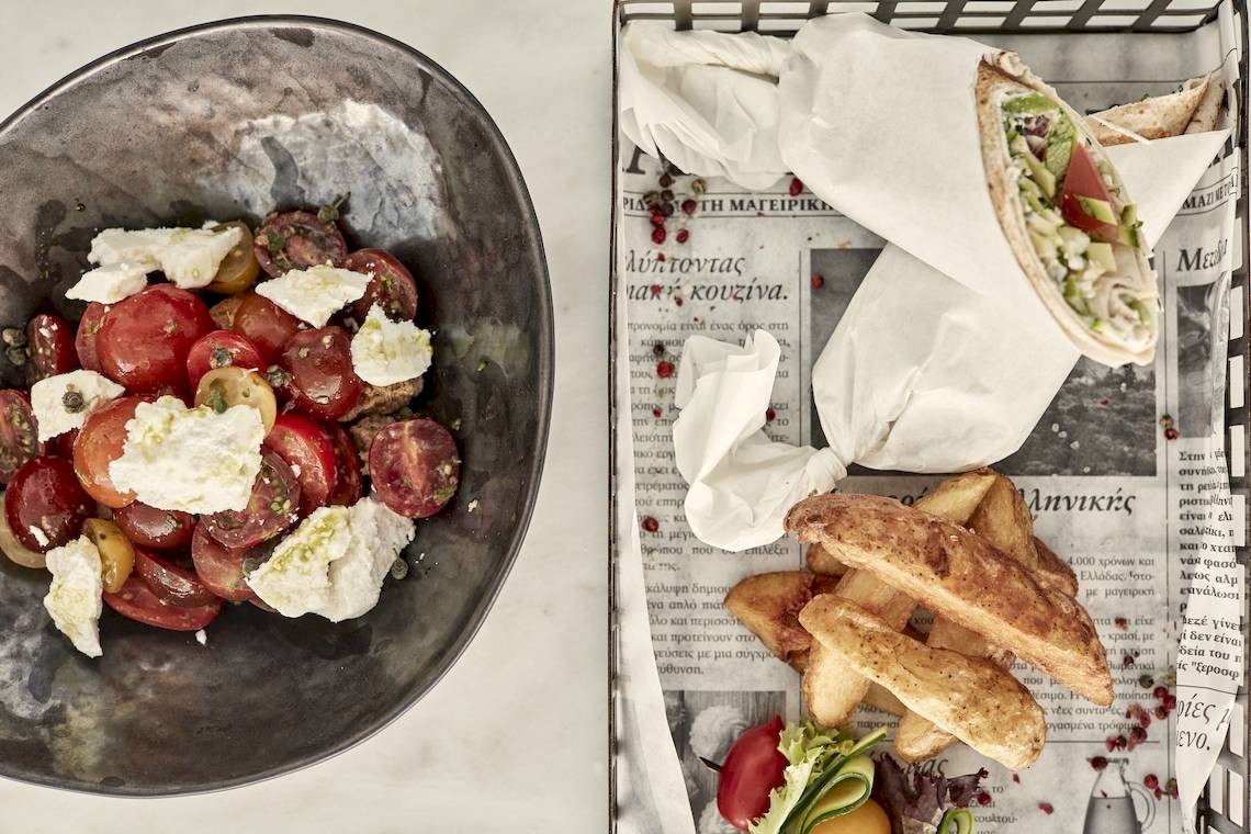 Ein Bild von einem Salat aus Tomaten und Feta in einer Schüssel und Pommes auf einer Zeitung.