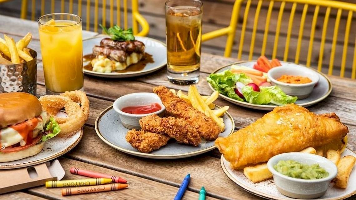 Essen und Getränke auf einem Holztisch im Tower Hotel, London. Darunter verschiedene Gerichte, Burger, Pommes, Salat und Getränke.