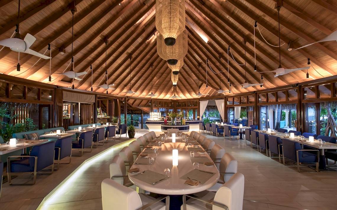 Innenansicht des Restaurants im Constance Ephelia Mahe Seychelles Hotel mit langen Tischen und Stühlen, hängenden Lampen und Holzdecken.
