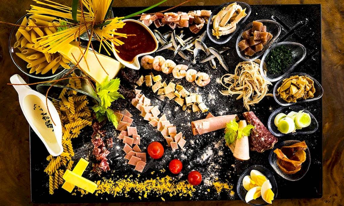 Frühstücksbuffet mit Pasta, Fisch, Schinken und Gemüse auf einem schwarzen Teller.
