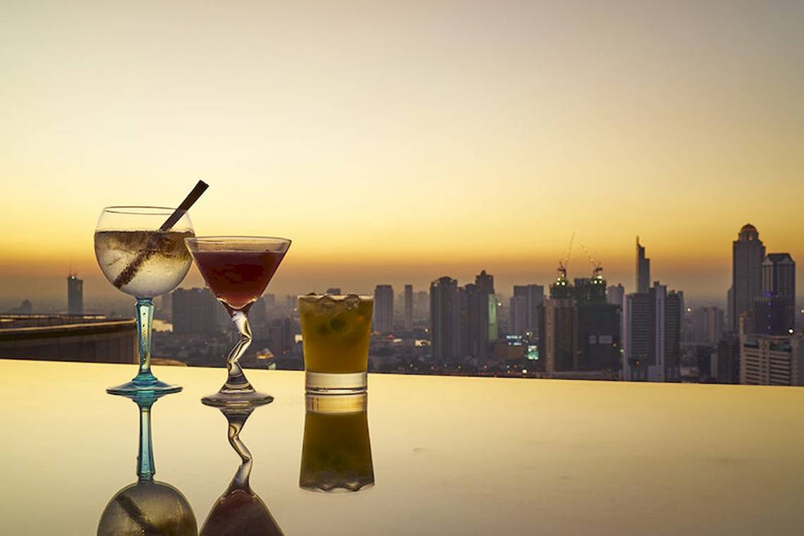Drei Gläser mit Getränken auf einer Terrasse mit Blick auf die Skyline einer Stadt.