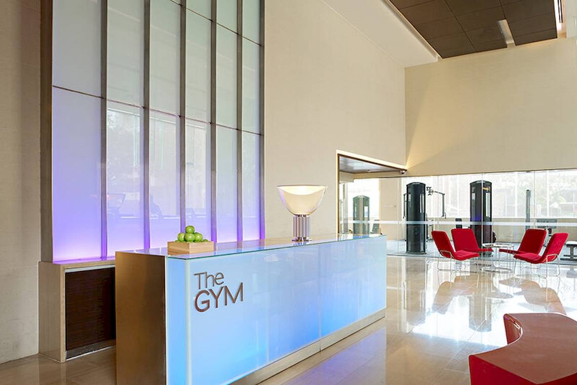 Lobby des Fitnessstudios im JC Kevin Sathorn Bangkok Hotel mit The Gym Schriftzug an der Theke und roten Stühlen.