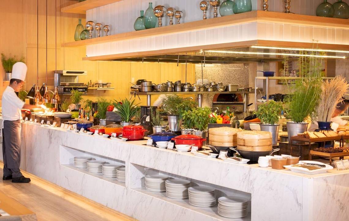 Restaurant im AVANI+ Riverside Bangkok mit Buffet und Köchen, die Speisen zubereiten.