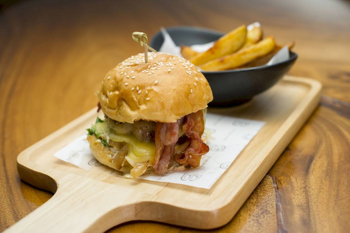 Ein Burger mit Speck und Pommes auf einem Holztablett auf einem braunen Tisch.