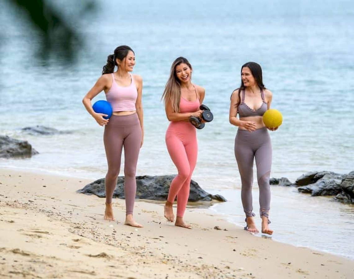 Drei Frauen am Strand mit Sportgeräten, Wasser im Hintergrund.