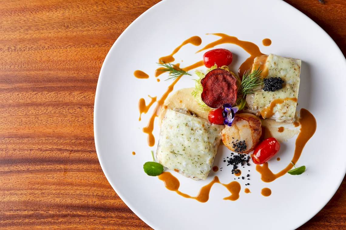 Ein Gericht auf einem weißen Teller auf einem Holztisch, mit Fisch, Tomaten und Kaviar garniert.