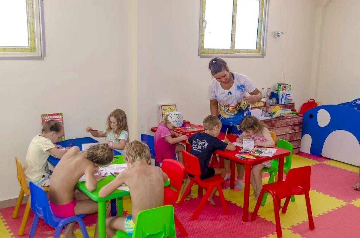 Gruppe von Kindern, die in einem farbenfrohen Raum um Tische herum sitzen und malen, während eine Frau ihnen hilft.