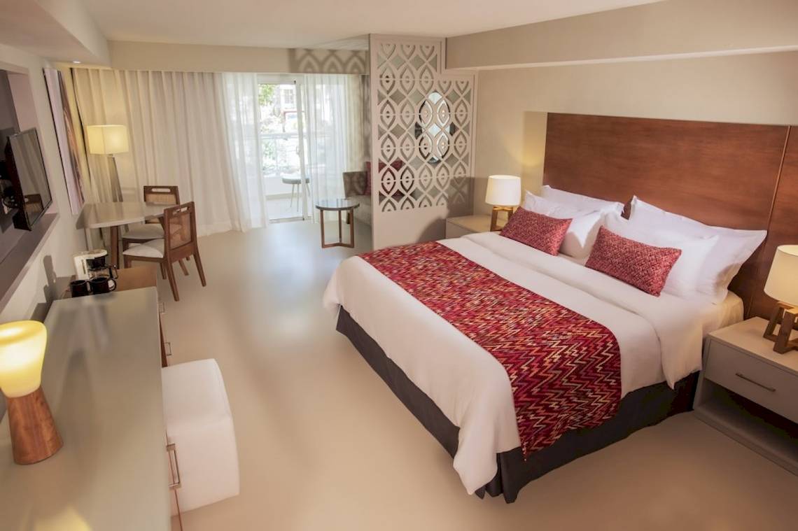 Innenansicht eines Hotelzimmers im Emotions By Hodelpa Puerto Plata mit Doppelbett, Tisch, Stühlen und Lampen.