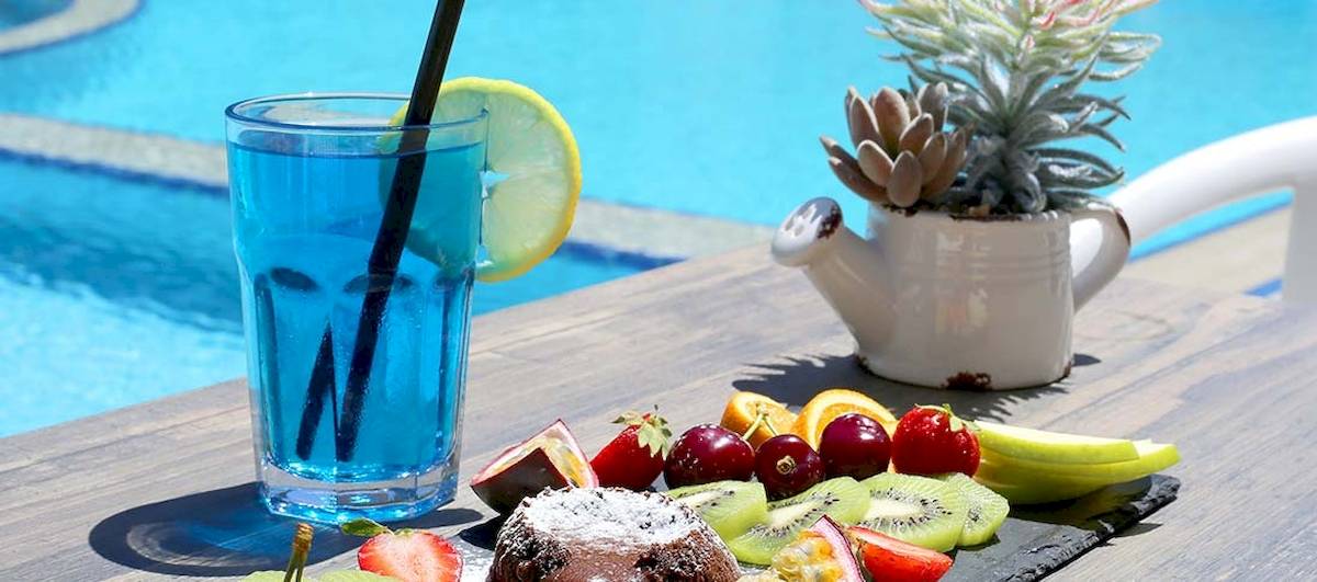 Dyrrah Hotel Poolseite mit einem Glas blauem Getränk, einer Zitronenscheibe, einer Obstplatte und einem Topf mit Sukkulenten neben dem Pool.