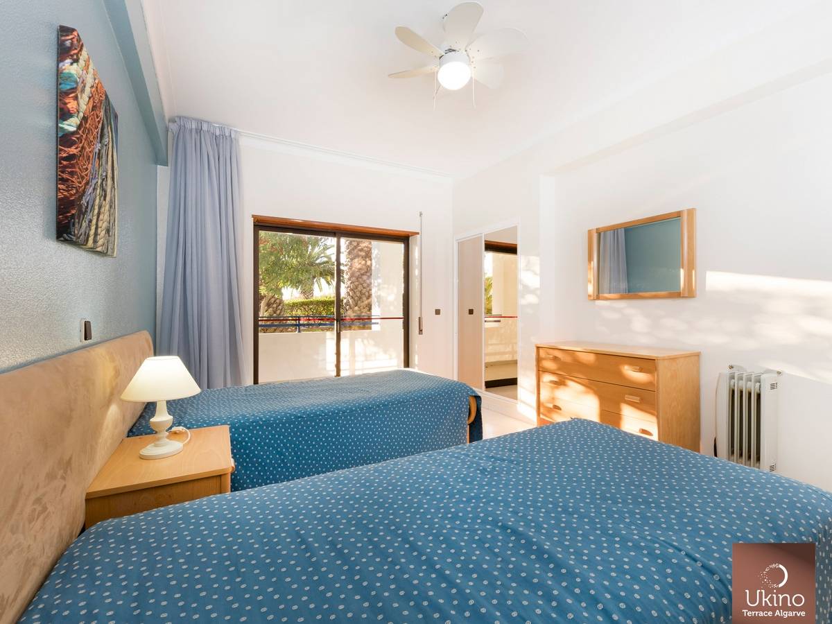Innenansicht eines Doppelzimmers im Ukino Terrace Algarve Hotel mit zwei Betten, Nachttischen, Deckenventilator und Fenstern.